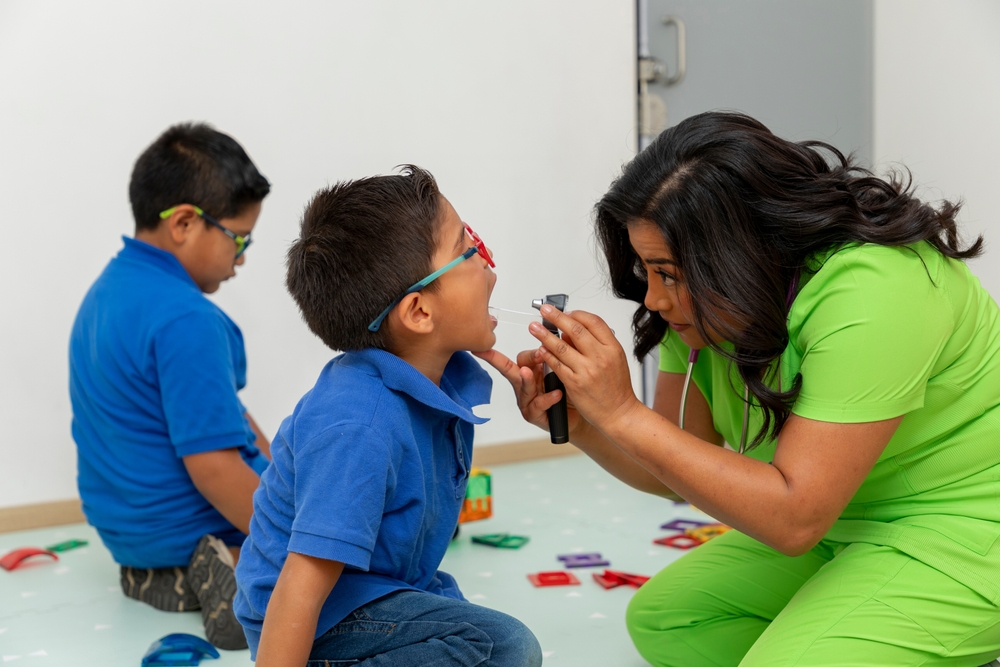 Mexican pediatrician examining a child's mouth with an otoscope inside her pediatric office