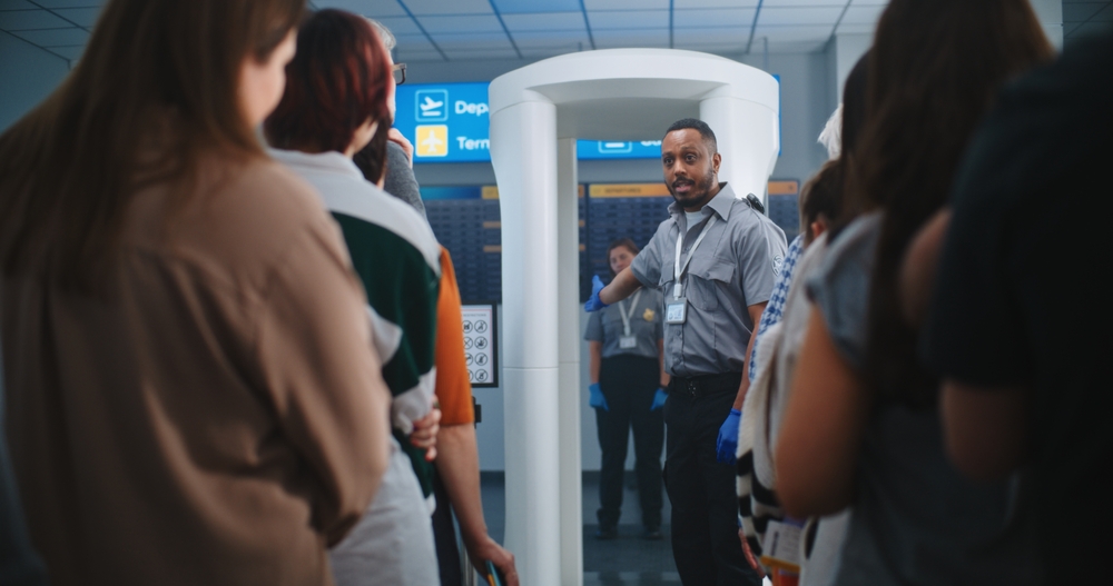 Airport Security Checkpoint: African Security Officer educates various people, passengers about Metal Detector Scanner Gates, TSA inspection procedures. A line of travelers on the move.
