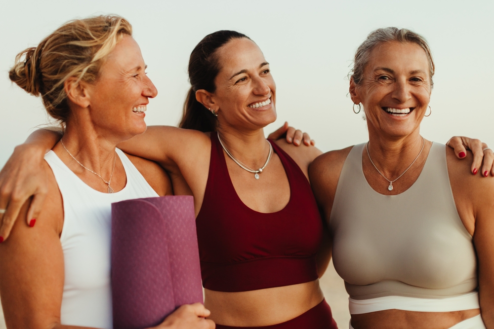 Three mature women share smiles and a bond of friendship while wearing yoga attire outdoors. The camaraderie and joy of their interaction reflect the essence of togetherness and wellbeing.