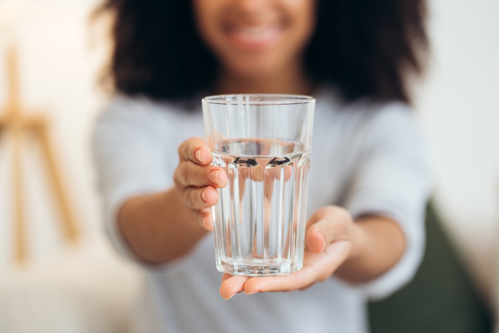 Smiling woman holding a glass of fresh water, promoting hydration and a healthy lifestyle while enjoying a moment of wellbeing at home