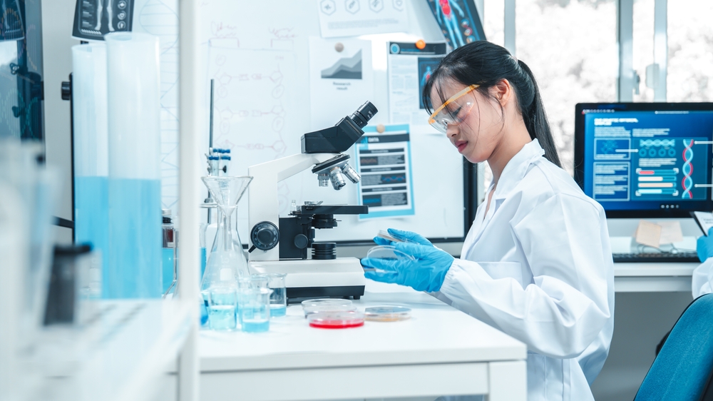 Asian female scientist analyzing microbial growth on petri dishes in modern laboratory. Laboratory research on fungal and bacterial cultures, with microscope and scientific data on screen.