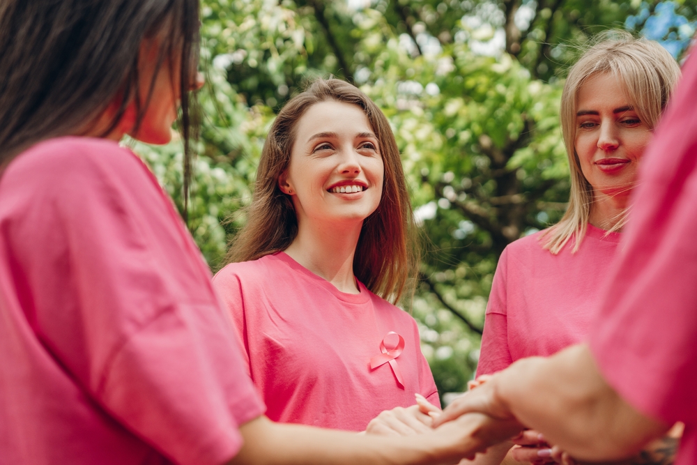 Volunteers wearing pink t-shirts with pink ribbons are holding hands, supporting breast cancer awareness, offering hope and unity in the fight against the disease. Health care concept