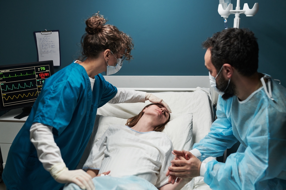 Caucasian young adult woman lying in hospital bed receiving medical attention from Caucasian female nurse and Caucasian male doctor, nurse checking forehead while doctor holding patient hand