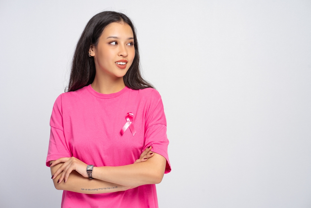 Smiling young woman wearing pink t-shirt with crossed arms and pink ribbon is looking on the side, showing support for breast cancer awareness, prevention and research