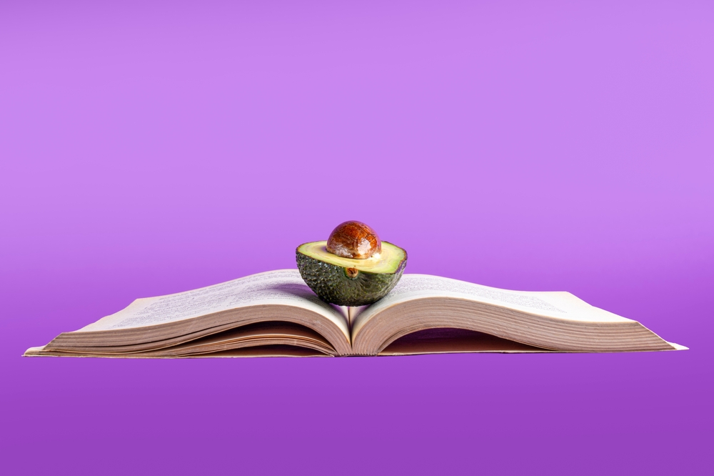Studio shot of a fresh avocado half with pit balanced on an open book against a purple background, representing healthy brain food for students.