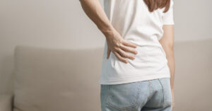 "Person wearing a white shirt and blue jeans holding their lower back, indicating back pain or discomfort.