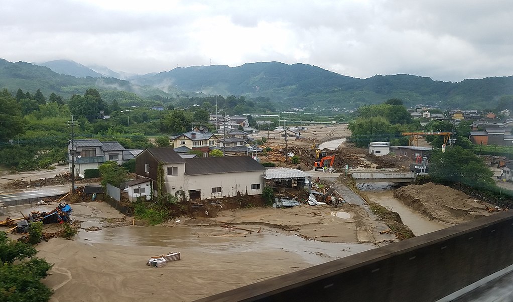 Flood-damaged Japanese town in a mountain valley, with mud-covered debris scattered around homes and a swollen brown river running through the wreckage.