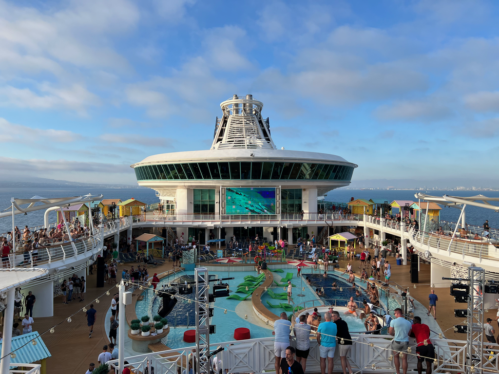 Passengers gather around a large pool deck on a cruise ship, with green inflatable loungers floating in the water and colorful beach cabanas lining both sides. A massive outdoor movie screen displays underwater footage beneath a distinctive circular observation lounge, while the coastline stretches across the horizon under a blue sky.