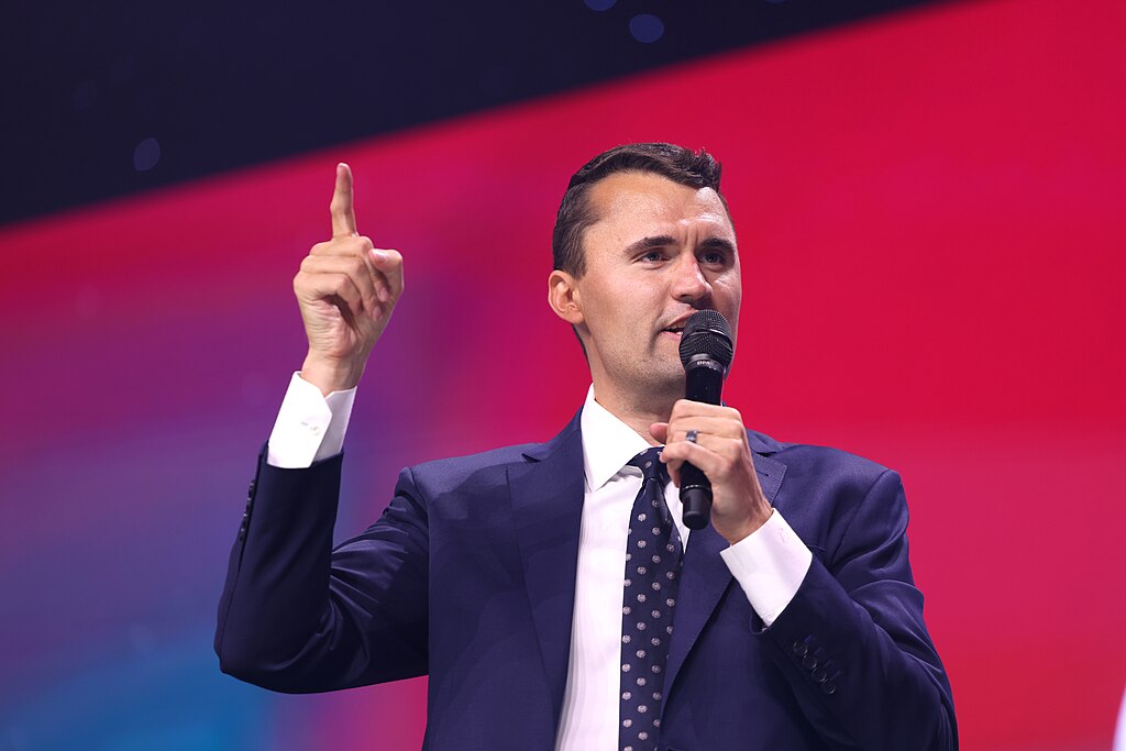 Charlie Kirk speaks at an event, holding a microphone in one hand while raising his index finger on the other. He wears a navy blue suit with a white dress shirt and dark patterned tie against a vibrant red and magenta gradient background.