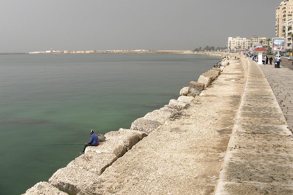 A fisherman sits on Alexandria's concrete seawall as it curves into the Mediterranean, with the Egyptian city's skyline visible through the haze across the harbor.