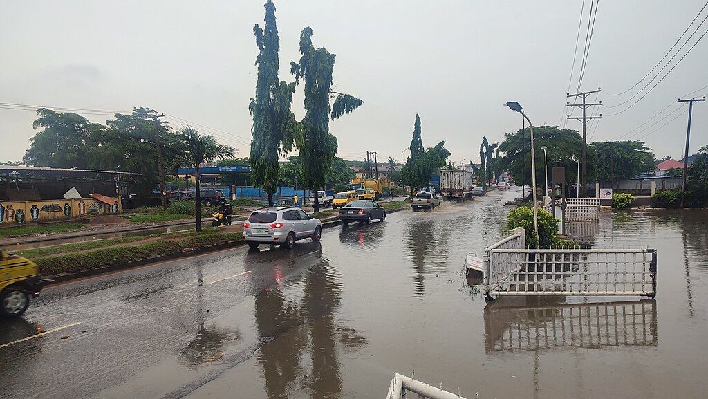 Vehicles navigate a flooded intersection under overcast skies, with standing water pooling across the road and a metal barrier partially submerged on the right.