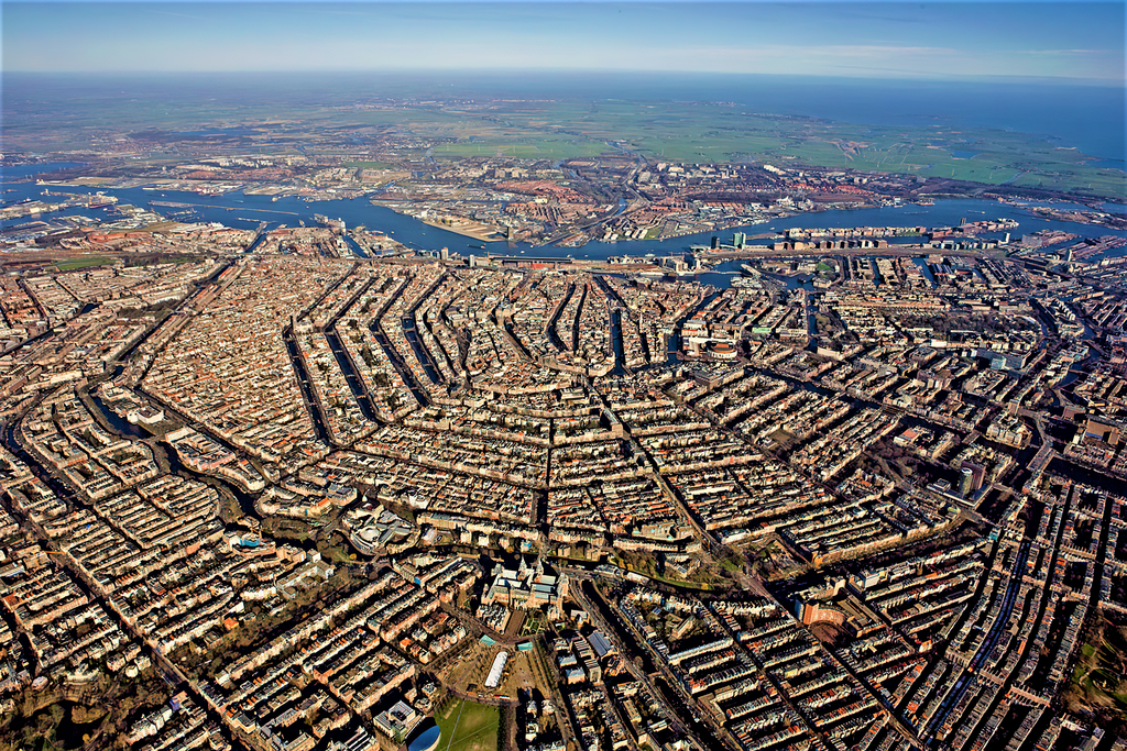 Aerial view of Amsterdam showing its distinctive semicircular canal ring pattern radiating outward from the historic center, with the IJ waterway visible along the northern edge.