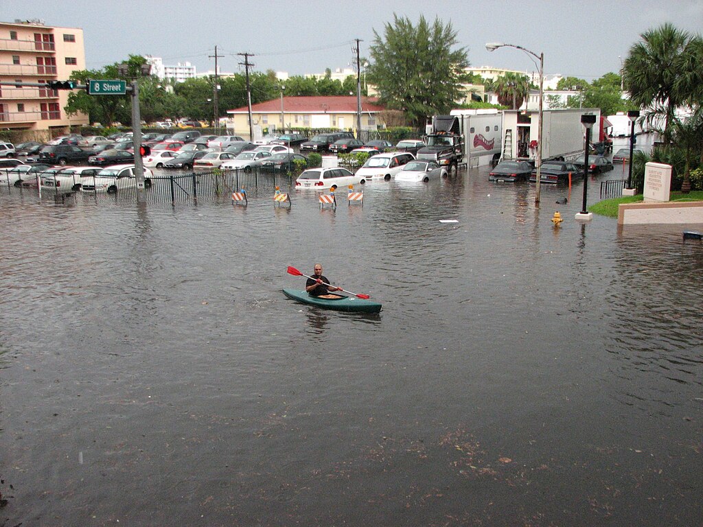 A kayaker paddling through a flooded city intersection, with water reaching car door height in a parking lot and street signs marking the roads now submerged.
