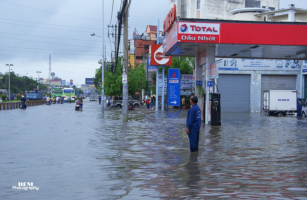 A man standing calf-deep in floodwater outside a gas station, with motorbikes and shops lining the submerged street behind him.