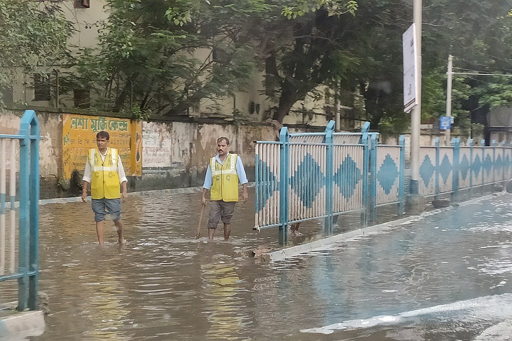 Two workers in yellow safety vests wade through knee-deep floodwater along a city sidewalk, with a blue metal railing separating them from the submerged street.