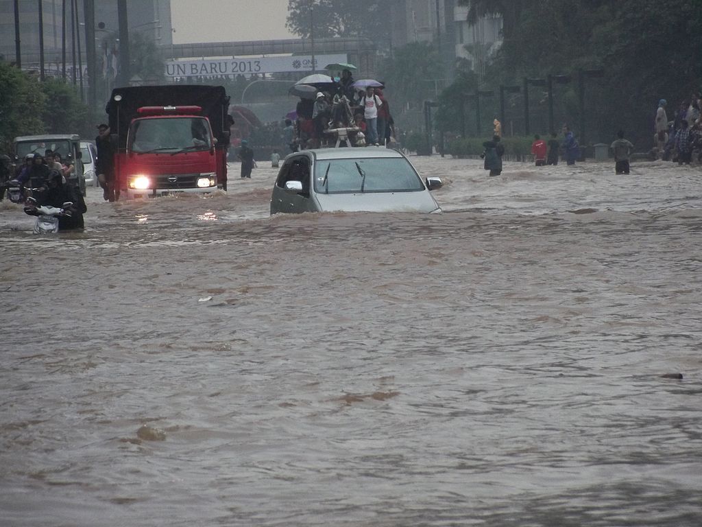 Vehicles stranded on a flooded urban street, with a white sedan submerged past its doors and a truck pushing through water while people gather on higher ground in the background.