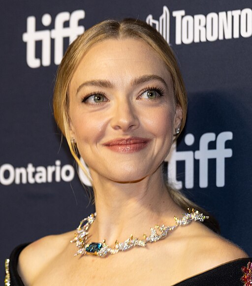 Amanda Seyfried at the Toronto International Film Festival, her blonde hair pulled back in a sleek style. She wears an elaborate statement necklace featuring blue gemstones and intricate metalwork, smiling warmly while looking slightly off-camera. The TIFF logo is visible in the background.