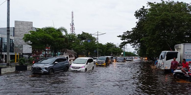 Cars push through a flooded commercial street, with water splashing around their tires and a cell tower visible against the gray sky.