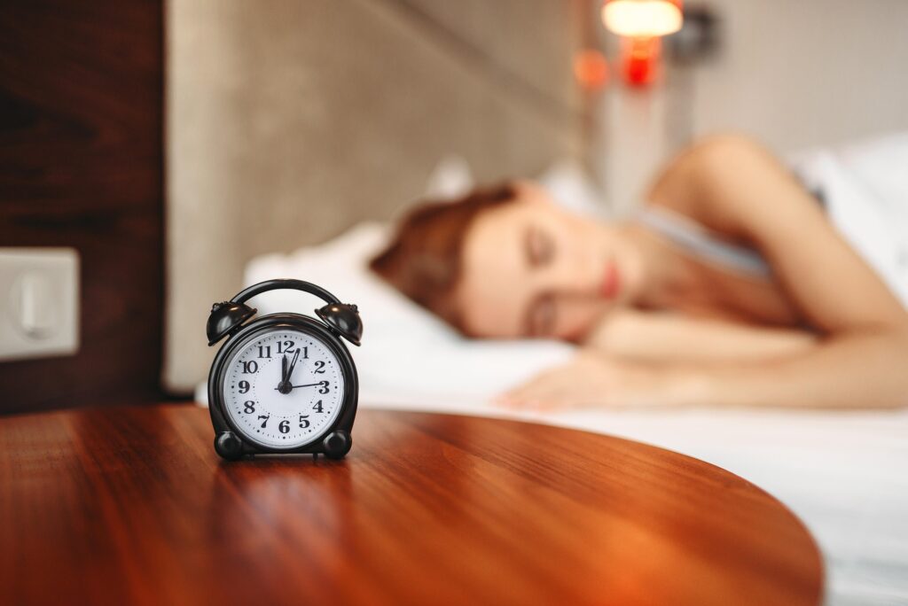 A black alarm clock sits on a wooden bedside table, its white face showing the time in sharp focus. Behind it, a woman with reddish hair sleeps on white bedding, her figure softly blurred. Warm light filters through the scene.