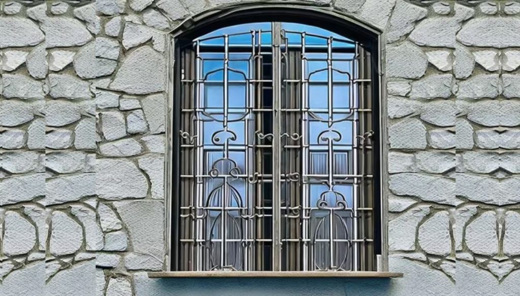 An intricately designed window grill on the window. A stone wall in the background. 