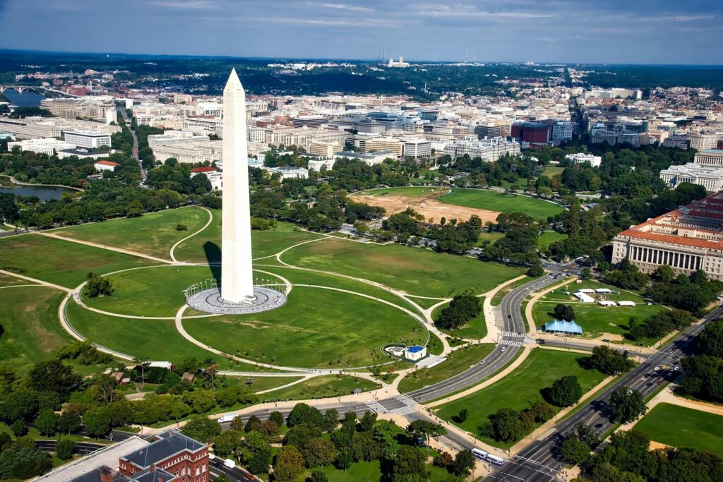 An aerial view of the Washington Monument rising from the National Mall, surrounded by green lawns and pathways, with the city stretching into the distance.