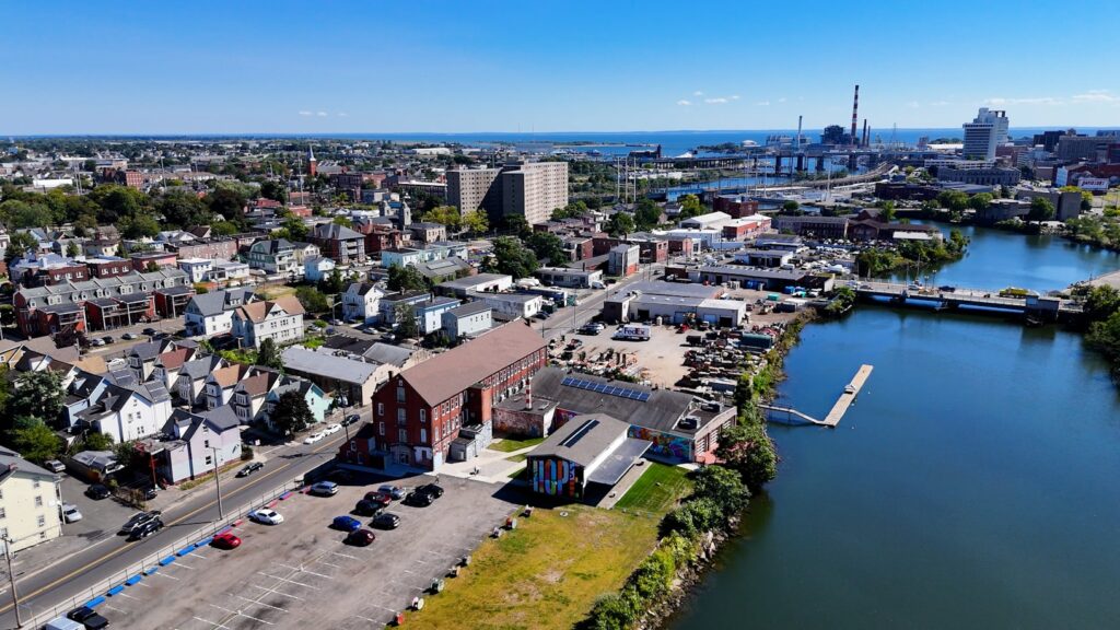 An aerial view of a Connecticut coastal town on a clear day, with brick buildings, a river inlet, and industrial structures visible in the distance.