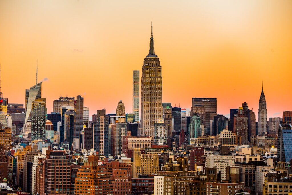 The Manhattan skyline at sunset, with the Empire State Building centered against an orange sky and surrounded by densely packed skyscrapers.