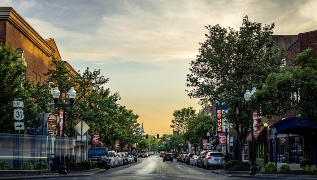 A tree-lined main street in a small town at sunset, with parked cars along both sides, neon signs on storefronts, and a golden sky glowing at the end of the road.