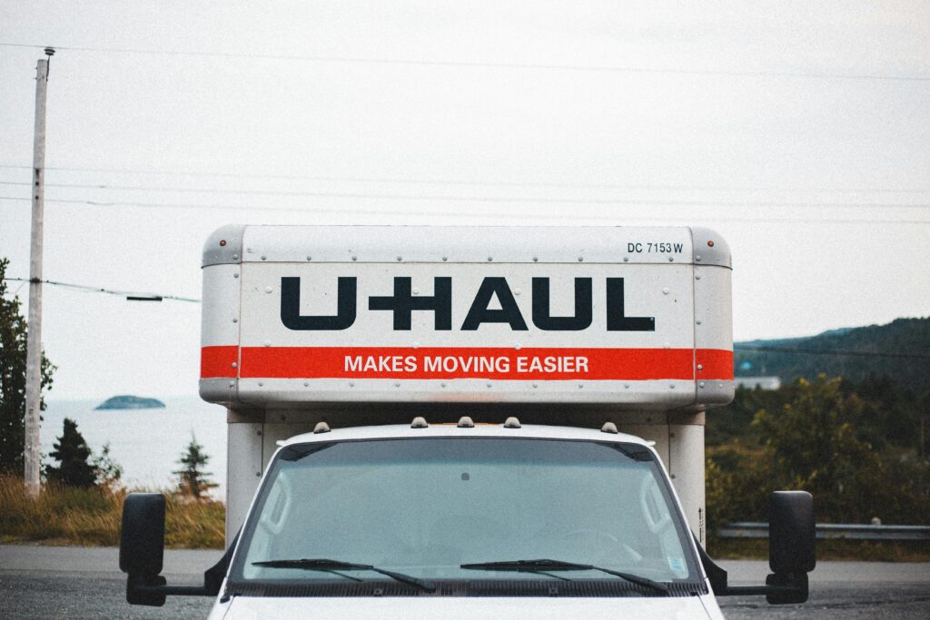 The front of a white U-Haul moving truck displaying the company's orange and red logo and the slogan "Makes Moving Easier," parked with a hilly, overcast landscape behind it.