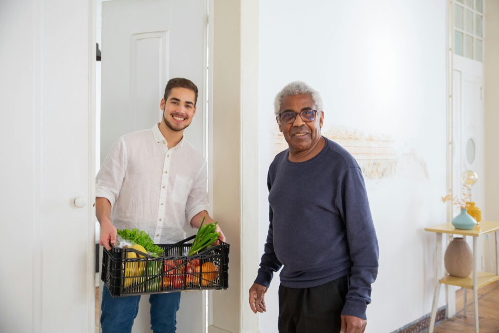 A young man holding a crate of fresh vegetables stands in a doorway while an older man in a blue sweater and glasses smiles beside him.