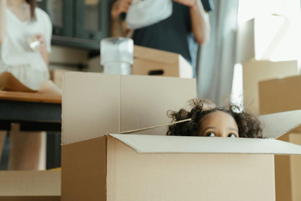 A young child with curly hair peeks out from inside a cardboard moving box, only their eyes and the top of their head visible above the edge. Adults move around in the soft-focus background among other packed boxes.