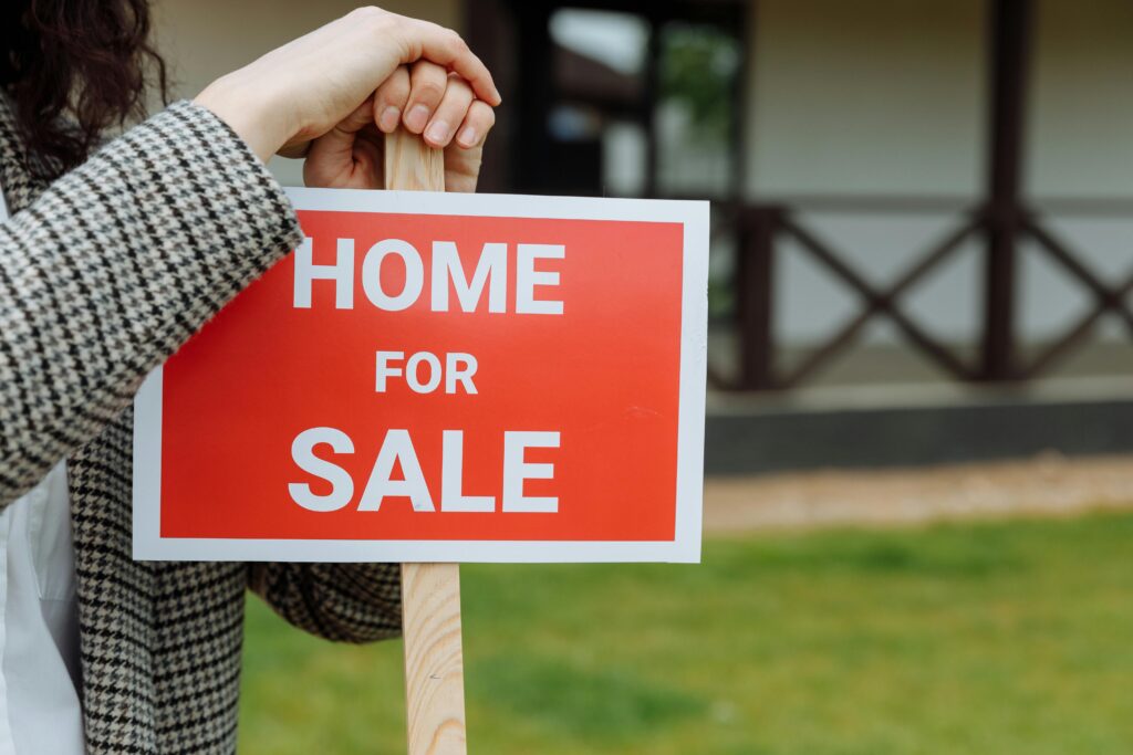 A person in a gray plaid jacket grips the wooden post of a red "Home For Sale" sign, with a house and lawn blurred in the background.