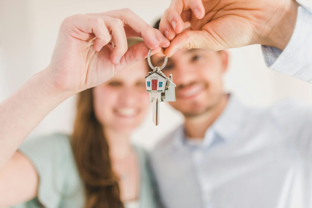 A smiling couple, blurred in the background, holds up a set of house keys with a house-shaped keychain toward the camera.