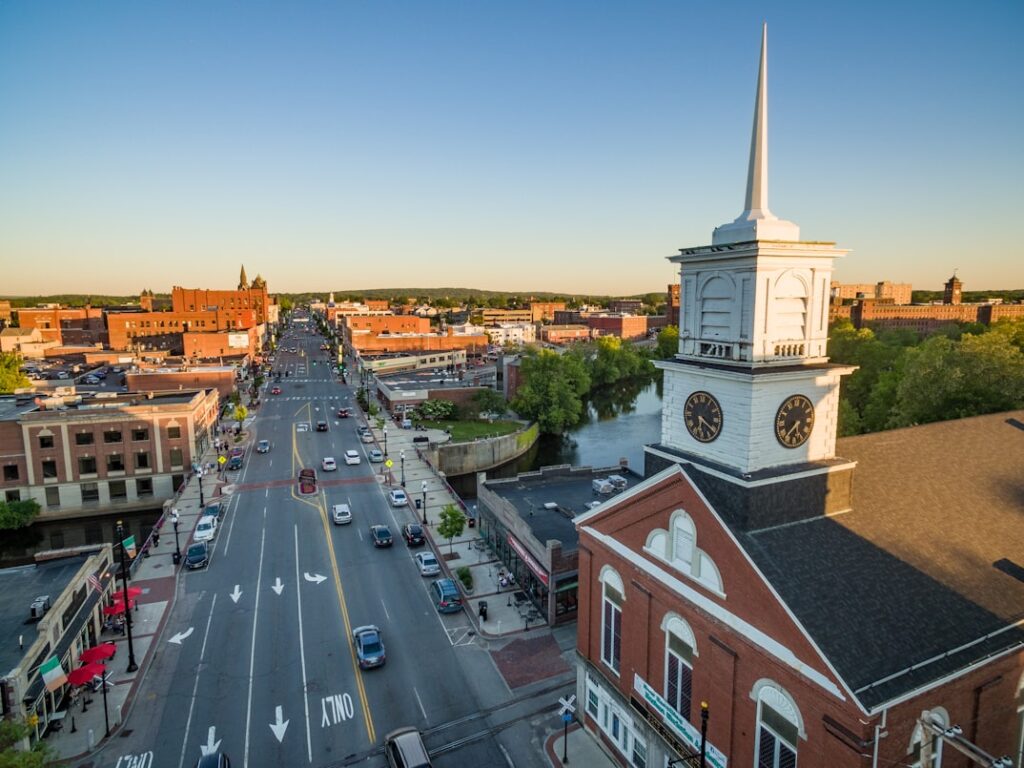 A wide main street runs through a New Hampshire town at golden hour, with a white church steeple rising beside brick storefronts and low hills in the background.