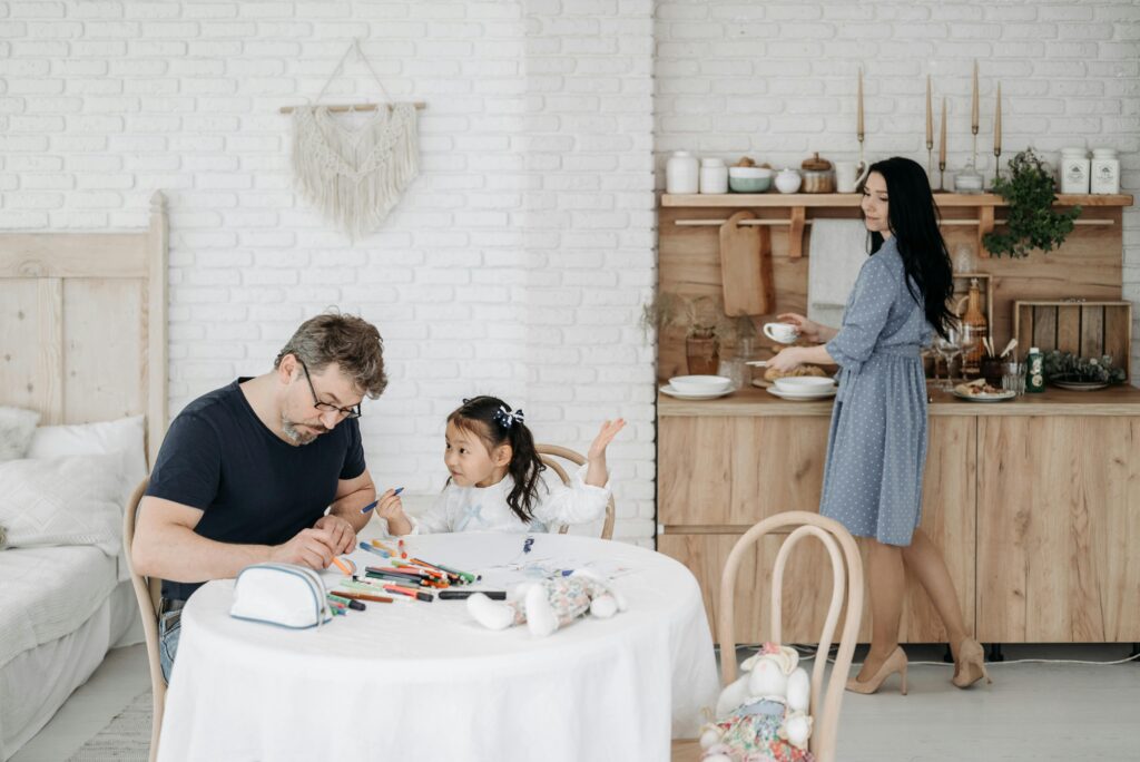 A father and young daughter sit at a table doing crafts together while a woman in a blue dress carries dishes in the background of a bright kitchen.