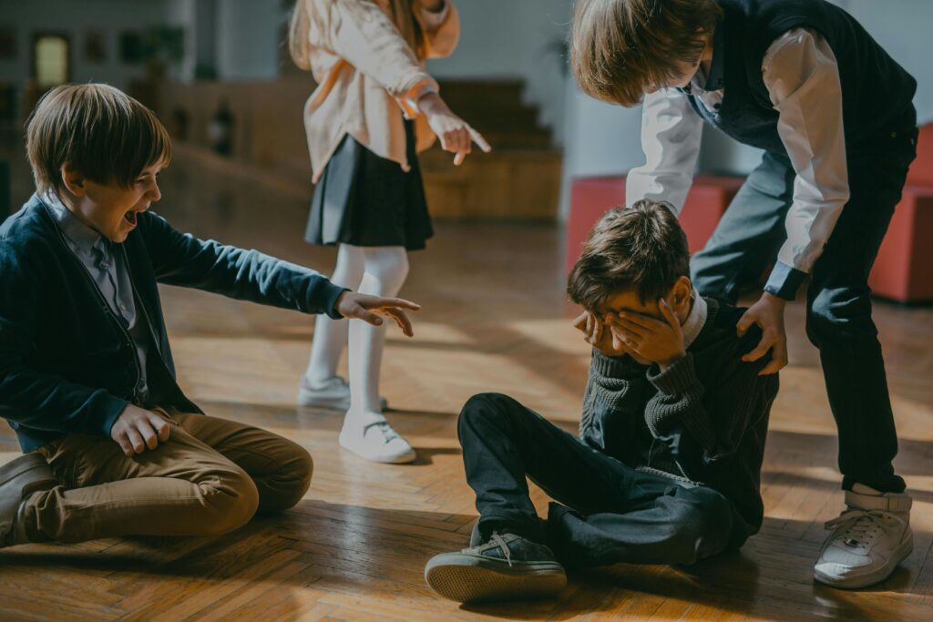 Children in school uniforms on a wooden floor, with one boy sitting and covering his face while three other children point and laugh at him in a bullying scene.
