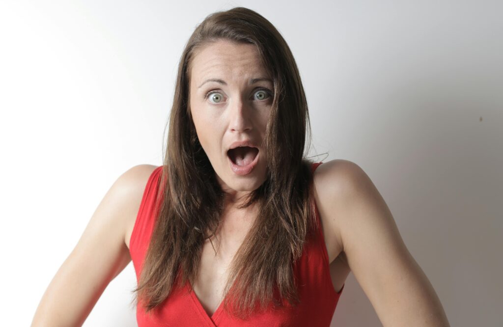 Woman with long brown hair wearing a red V-neck top, mouth wide open in an exaggerated expression of shock against a plain white background.