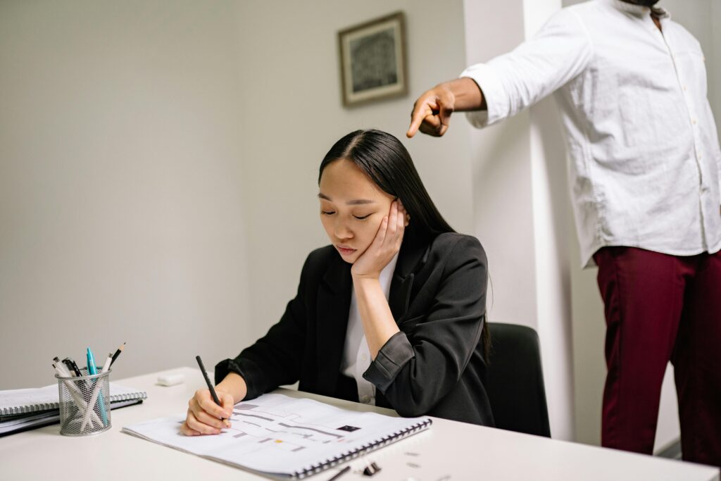 Woman in a black blazer sits at an office desk writing, resting her cheek on her hand with a dejected expression while a standing coworker points down at her critically.