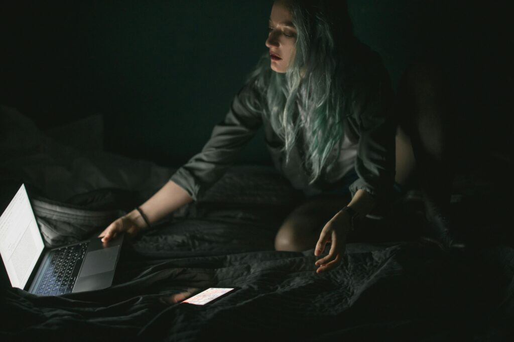 A woman with long gray-blue hair sits cross-legged on a bed in darkness, her face lit by the glow of an open laptop while she holds her phone.