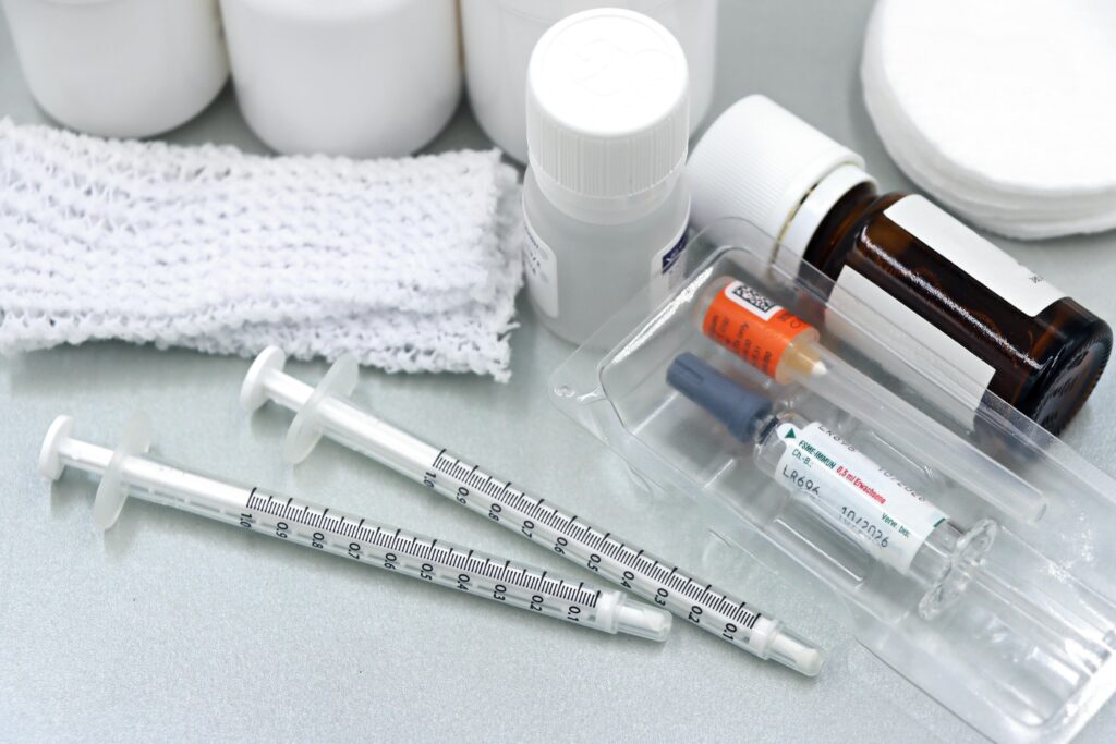 Three small syringes lie on a gray surface alongside vaccine vials, a brown glass medicine bottle, white gauze, and plastic caps in orange, blue, and gray.