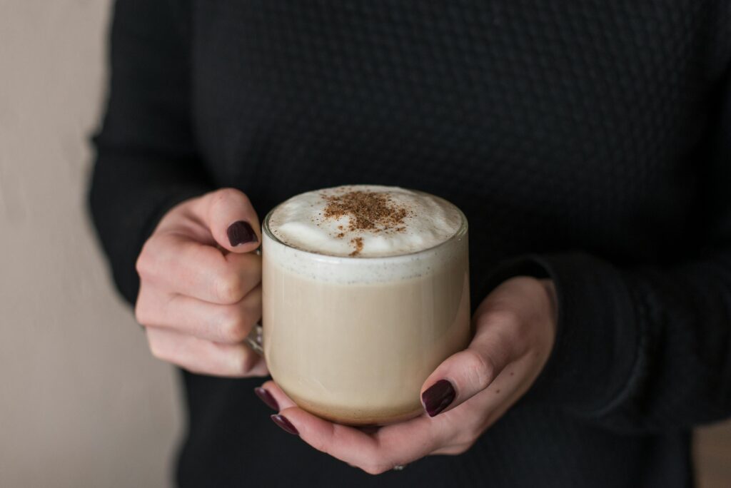 Hands with dark nail polish cradling a glass mug of foamy latte.