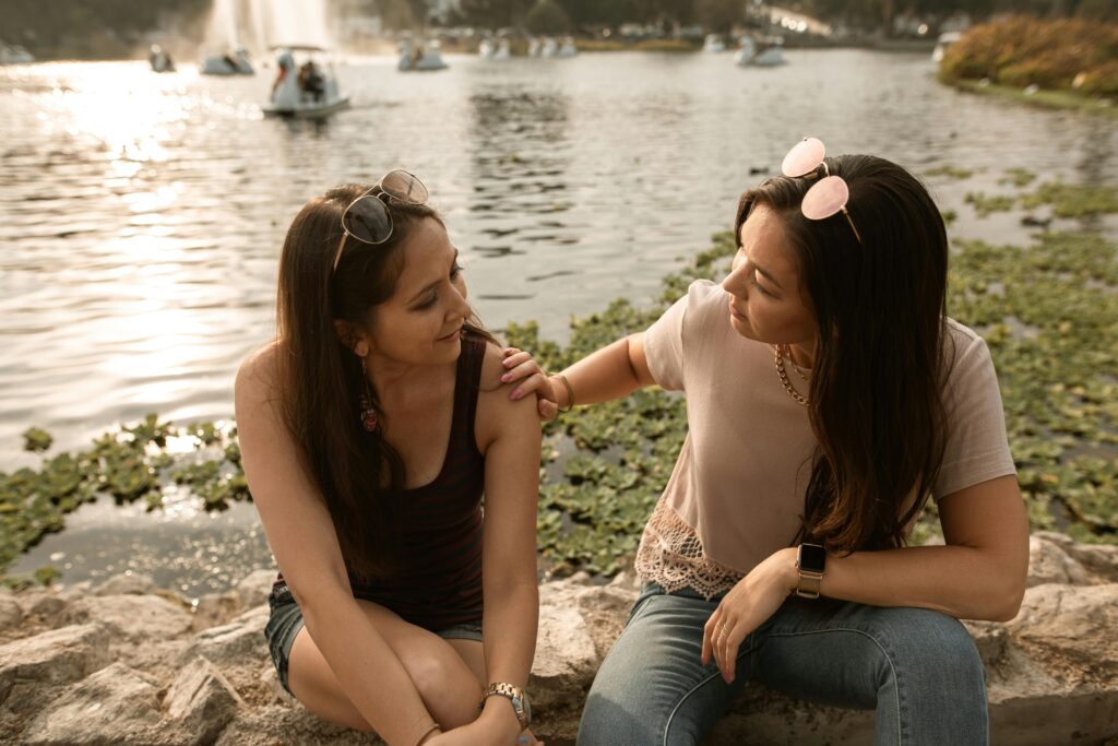 Two women sitting by a lake at golden hour, one resting her hand on the other's shoulder mid-conversation.