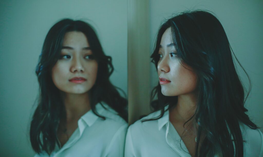 A woman in a white button-down shirt looking at her own reflection, both faces visible in soft blue-green light.