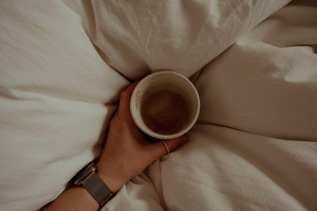 A person's hands cradle a mug of dark tea or coffee while resting in bed, surrounded by rumpled white linens. They wear a silver watch on their wrist. The overhead shot creates an intimate, cozy morning atmosphere.