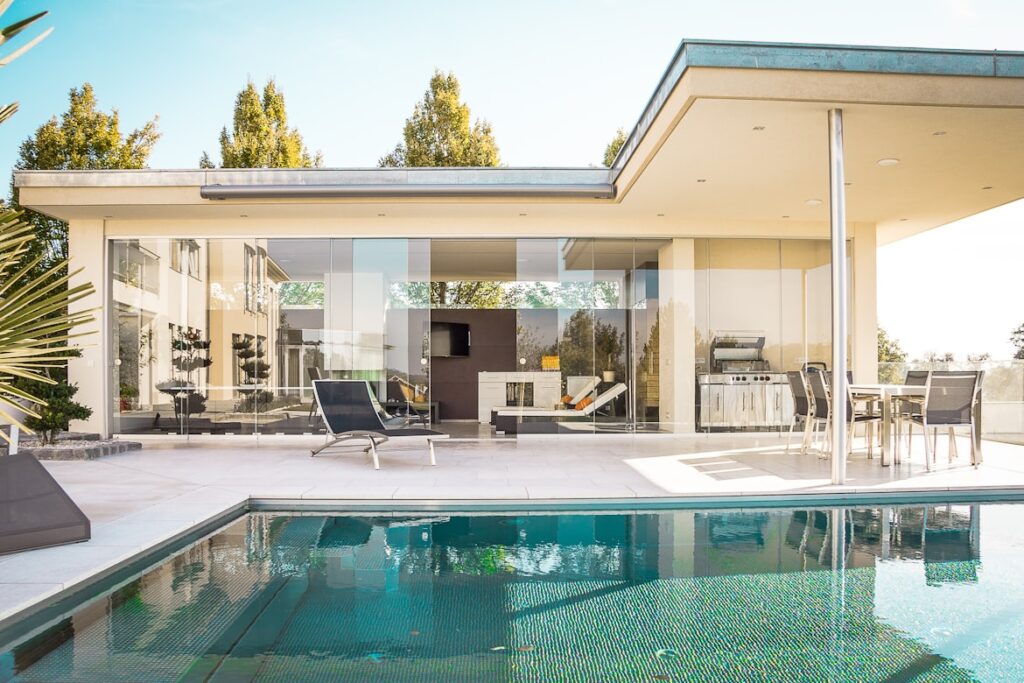 A sleek single-story home with floor-to-ceiling glass walls opening onto a patio and turquoise swimming pool, with modern outdoor furniture arranged in the sun.