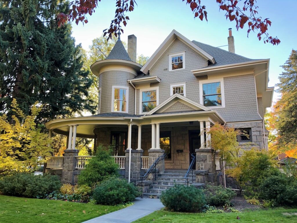 A gray Victorian home with a rounded turret, covered front porch with white columns, and decorative trim, surrounded by trees with autumn leaves.