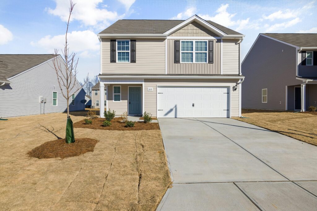 A beige two-story suburban home with an attached garage, small front yard with fresh mulch, and a newly planted tree in a cookie-cutter neighborhood.