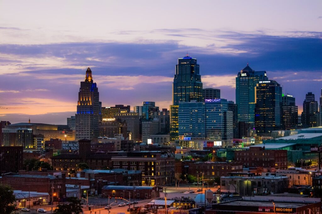 The Kansas City skyline glows at twilight, with illuminated high-rises and the art deco Power and Light Building standing against a purple and orange sky.