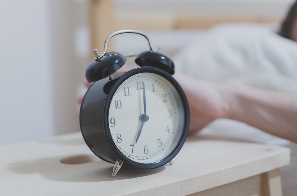 A black twin-bell alarm clock on a wooden nightstand shows 7 o'clock as a hand reaches toward it from bed.