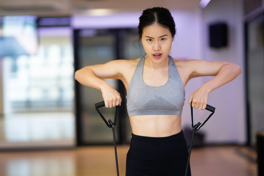 Young beautiful Asian women workout at the gym indoor. She is using the elastic band to stretch her arms and legs. She is wearing gym clothes grey bra outfit and get sweaty.